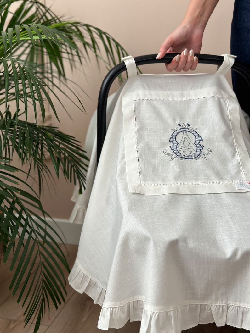 May include: White linen stroller cover with a ruffled edge and an embroidered monogram in the center. The monogram is a stylized letter "A" inside a circle.