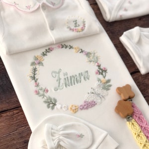 May include: A white baby outfit set with floral embroidery. The set includes a onesie, a sleep sack, a hat, and mittens. The sleep sack has the name "Z&uuml;mra" embroidered on it. The set also includes a wooden teether with a yellow and pink braided strap.