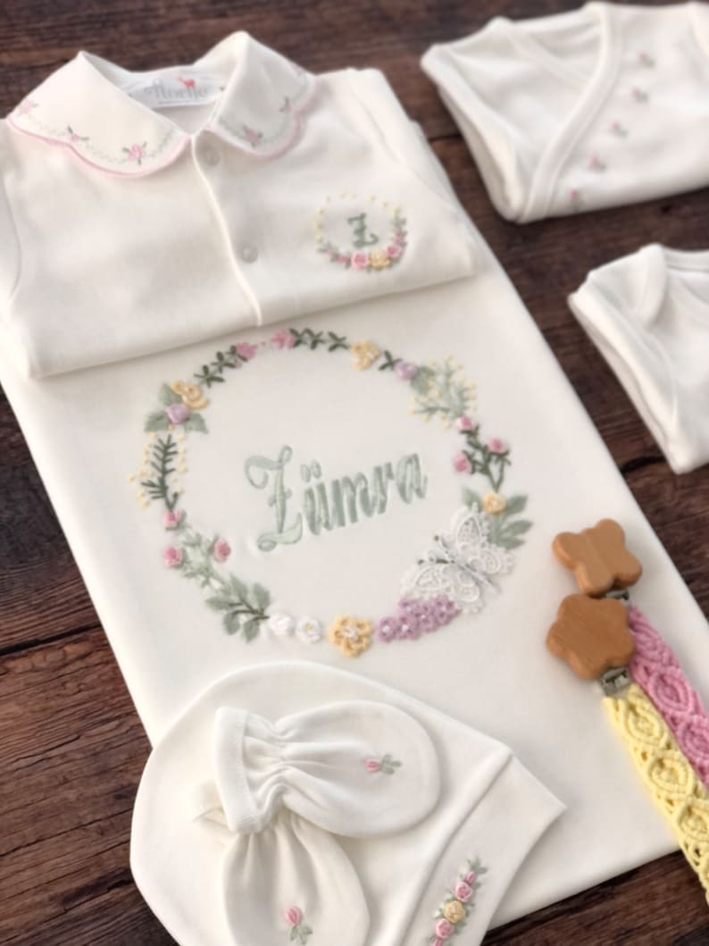 May include: A white baby outfit set with floral embroidery. The set includes a onesie, a sleep sack, a hat, and mittens. The sleep sack has the name "Z&uuml;mra" embroidered on it. The set also includes a wooden teether with a yellow and pink braided strap.