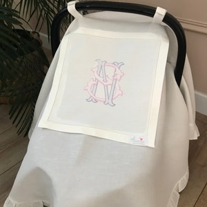 May include: White car seat cover with a ruffled edge and a monogrammed initial "S" in pink and gray. The cover is made of linen and has a small pink butterfly logo with the word "florene" on it.