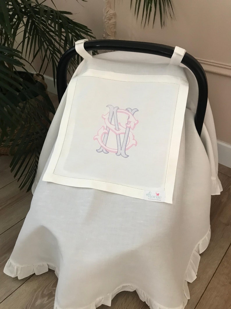 May include: White car seat cover with a ruffled edge and a monogrammed initial "S" in pink and gray. The cover is made of linen and has a small pink butterfly logo with the word "florene" on it.