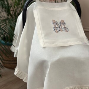 May include: White stroller cover with a ruffled edge and a monogrammed initial "M" in a brown and blue embroidery design.