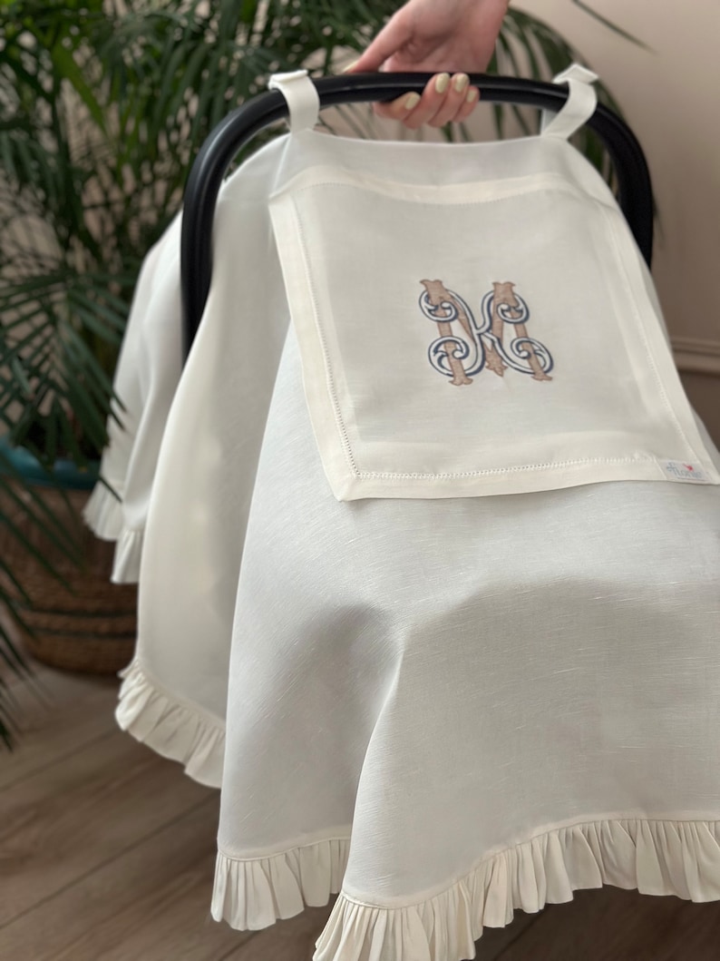 May include: White stroller cover with a ruffled edge and a monogrammed initial "M" in a brown and blue embroidery design.