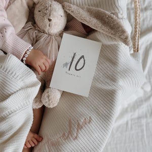 May include: A beige plush rabbit toy with a tulle skirt, a white milestone card featuring the number 10 and the text "MONATE ALT" with a small elephant illustration, and a cream-colored knitted blanket embroidered with the name "Amelia".