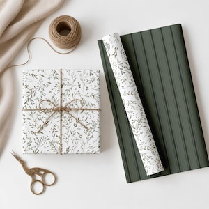 Könnte beinhalten: Ein quadratisches Geschenkpaket in weißem Papier mit grünem Blatt- und rotem Beerenmuster, gebunden mit brauner Schnur. Daneben liegen eine Rolle des gleichen Papiers, ein Bogen dunkelgrün gestreiftes Papier, eine Garnrolle und eine goldene Schere.