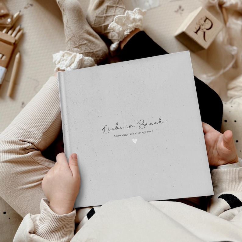 May include: A gray, textured journal with the text "Liebe im Bauch" and "Schwangerschaftstagebuch" in black script. A small white heart is below the text.
