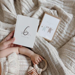 May include: A white rectangular milestone card featuring the number 6 and the text "MONATE ALT" in a dark gray font, decorated with small botanical illustrations. A second card with a graphic of hanging baby clothes is visible in the background.