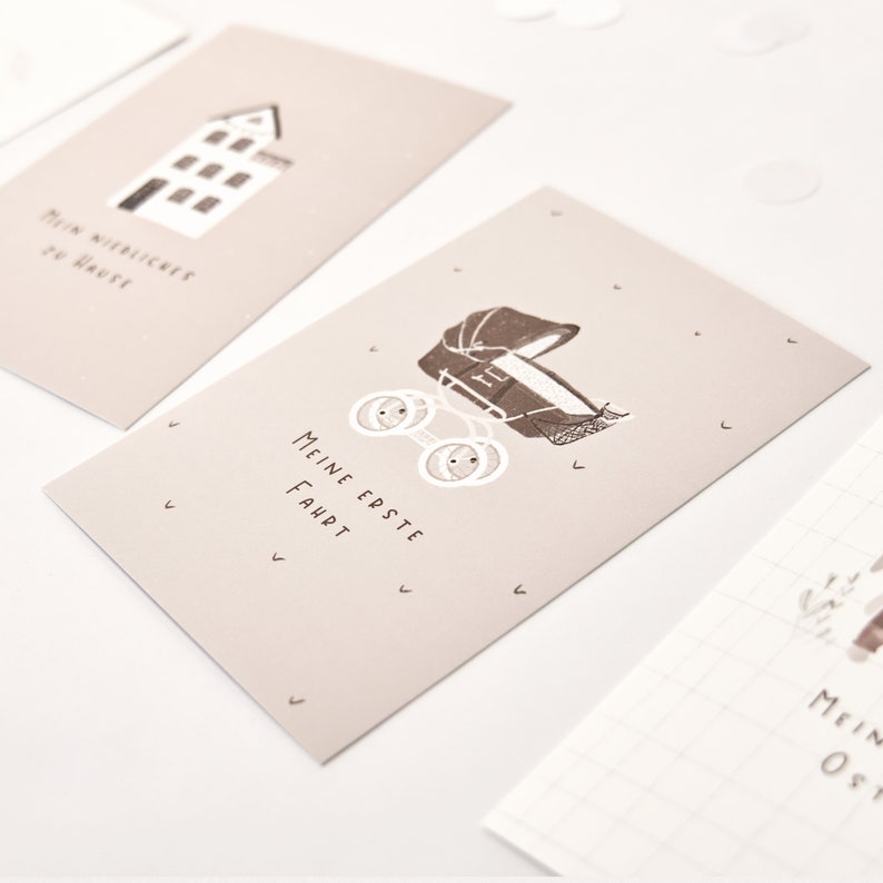 May include: A rectangular beige card featuring a dark brown illustration of a vintage baby carriage with four wheels. The text "MEINE ERSTE FAHRT" is printed in dark brown below the carriage. Small dark brown check marks are scattered around.