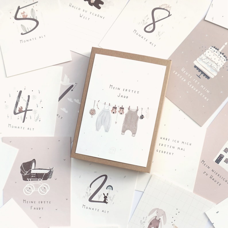 May include: A set of milestone cards for infants, featuring a central box labeled Mein erstes Jahr with illustrations of baby clothes on a clothesline, surrounded by various cards displaying numbers, illustrations of a cake, a pram, and German text.