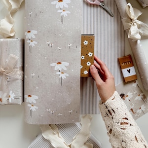 Op de afbeelding: Een set cadeauverpakkingsmaterialen bestaande uit een groot vel beige papier met een wit madeliefjespatroon, een kleine bruine doos met witte bloemen, een rol gestreept papier, een kleine rechthoekige doos en crèmekleurige linten.