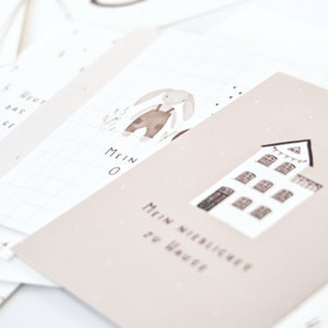 May include: A set of light beige and white cards featuring watercolor illustrations. One card displays a house with the text "MEIN NIEDLICHER ZU HAUSE" and another shows a rabbit in overalls with the text "MEIN O". The cards have a grid pattern.