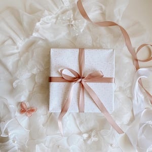 Könnte beinhalten: Eine quadratische Geschenkbox, eingewickelt in weißes Papier mit einem dezenten hellgrauen Blumenmuster, gebunden mit einer glatten roségoldenen Satin-Schleife. Die Box ist umgeben von weißem Rüschenstoff, einem kleinen roségoldenen Schmetterling und weißen Bändern.
