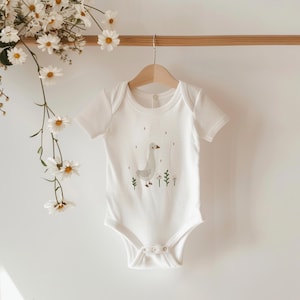 Könnte beinhalten: Weißer Baby-Strampler mit kurzen Ärmeln und einem grauen Gänse-Motiv mit grünem Gras und weißen Blumen.