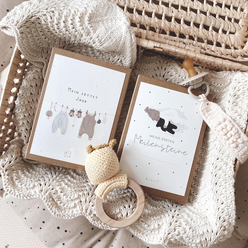 May include: Two rectangular cards with kraft paper borders featuring illustrations of baby clothes and a child, labeled Mein Erstes Jahr and Meine Ersten Meilensteine. Includes a beige crocheted rattle with a wooden ring and a pacifier with a crocheted strap.