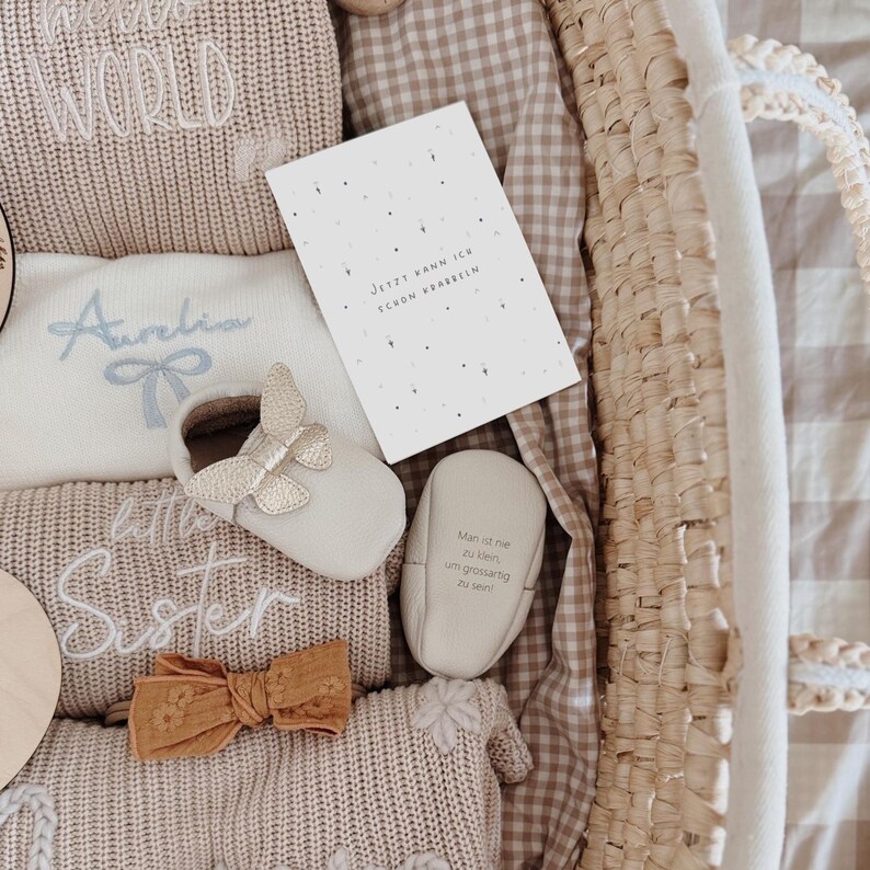 May include: A collection of baby items including a beige knit sweater with Hello World text, a white onesie with the name Aurelia and a blue bow, a mustard yellow hair bow, and cream-colored baby shoes with butterfly accents and German text.