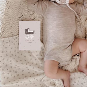 May include: A beige long-sleeved bodysuit and a light-colored crocheted bonnet. Beside the clothing is a rectangular card featuring a dark brown illustration of a vintage baby carriage and the German text Meine erste Fahrt.