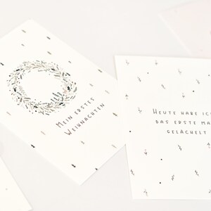 May include: Two white rectangular cards with black text and botanical illustrations. One card features a wreath and the text Mein Erstes Weihnachten. The other card displays the text Heute habe ich das erste Mal gel&auml;chelt with small floral accents.