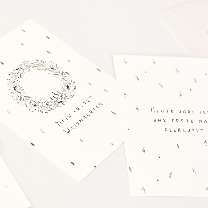 May include: Two white rectangular cards with black text and botanical illustrations. One card features a wreath and the text Mein Erstes Weihnachten. The other card displays the text Heute habe ich das erste Mal gel&auml;chelt with small floral accents.