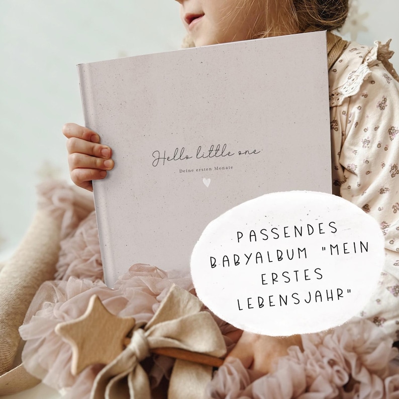 May include: A pink baby memory book with the text "Hello little one" and "Deine ersten Monate" on the cover. The book is being held by a child wearing a pink dress. The text "Passendes Babyalbum "Mein erstes Lebensjahr" is visible in the background.