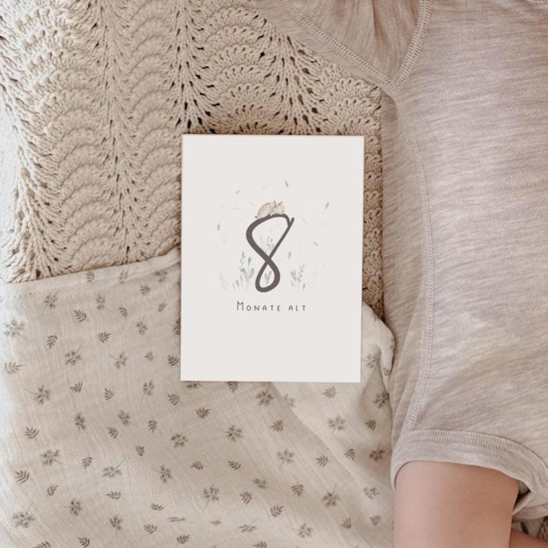 May include: A rectangular white milestone card featuring a large dark gray number 8, a small illustration of a sleeping animal, and the text "MONATE ALT" in dark gray. The card is positioned on a light beige fabric surface with a subtle pattern.