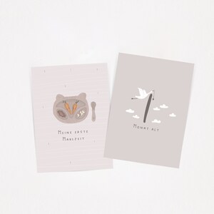 May include: Two rectangular milestone cards with light pink backgrounds. The left card features a bear-shaped plate with carrots and the text "Meine erste Mahlzeit". The right card shows a stork carrying a bundle, the number 1, and "Monat alt".