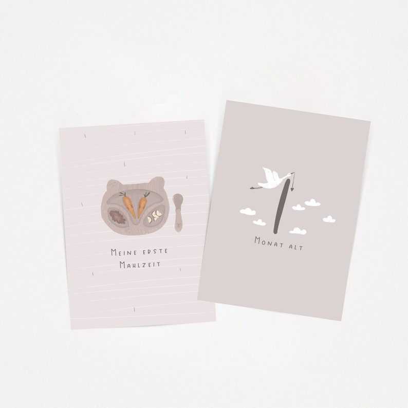 May include: Two rectangular milestone cards with light pink backgrounds. The left card features a bear-shaped plate with carrots and the text "Meine erste Mahlzeit". The right card shows a stork carrying a bundle, the number 1, and "Monat alt".