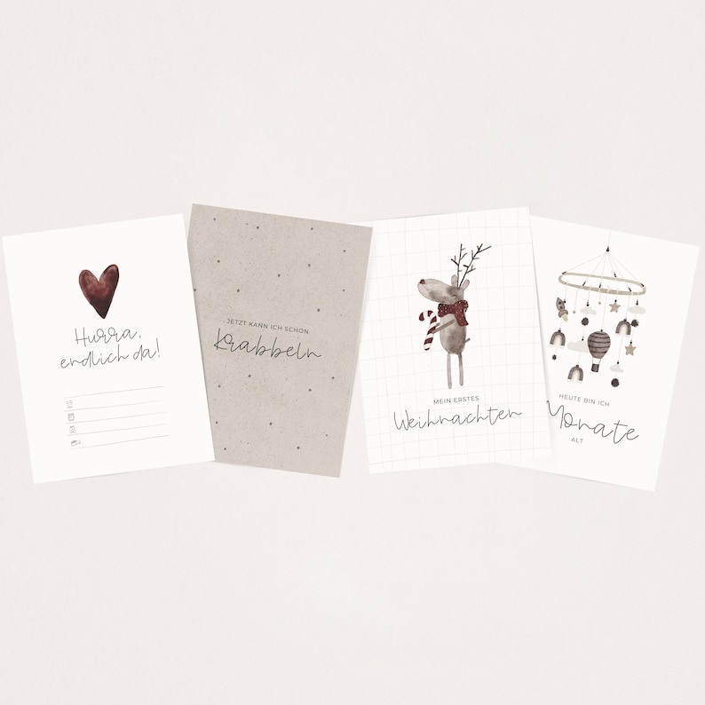 May include: Four printable cards with illustrations and text. The first card is white with a red heart and the text "Hurra. endlich da!" The second card is brown with the text "JETZT KANN ICH SCHON Krabbeln". The third card is white with a grid pattern and a reindeer illustration with the text "MEIN ERSTES Weihnachten". The fourth card is white with a mobile illustration and the text "HEUTE BIN ICH Monate ALT".