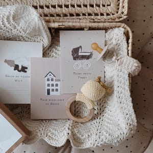 May include: A wicker basket containing a cream-colored knit blanket, a light-colored pacifier, a crochet rattle with a wooden ring, and three cards with illustrations and German text including "Hallo du sch&ouml;n Welt", "Meine erste Fahrt", and "Mein niedliches zu Hause".