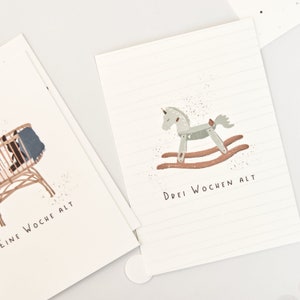 May include: Two white rectangular cards with thin horizontal lines. One card features a brown rocking horse illustration and the text "DREI WOCHEN ALT". The other card shows a brown cradle with a blue blanket and the text "EINE WOCHE ALT".