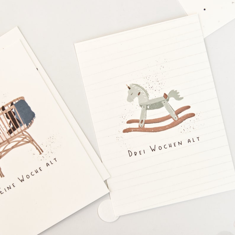 May include: Two white rectangular cards with thin horizontal lines. One card features a brown rocking horse illustration and the text "DREI WOCHEN ALT". The other card shows a brown cradle with a blue blanket and the text "EINE WOCHE ALT".