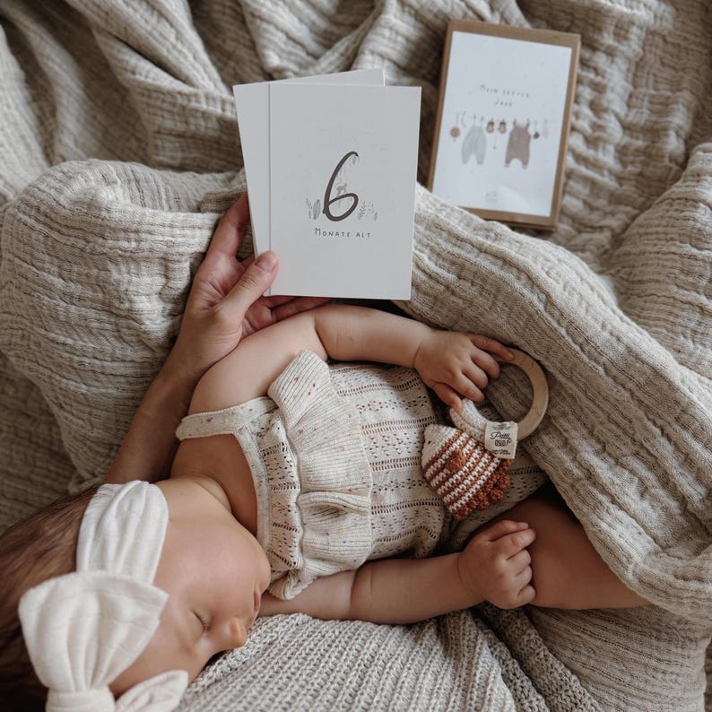May include: A white milestone card with the number 6 and the text 'MONATE ALT' printed in black, accompanied by a cream-colored knit romper, a matching headband, and a wooden ring rattle with a brown and white crocheted animal attachment.