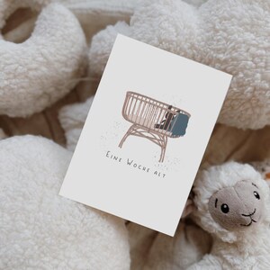 May include: A rectangular white card featuring a minimalist illustration of a light brown wicker bassinet with a small dark blue blanket inside. Below the illustration, the text reads "EINE WOCHE ALT" in a simple, dark gray sans-serif font.