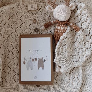 May include: A cream-colored knit cardigan with wooden buttons, a crocheted deer toy with a brown and white patterned body, and a rectangular box featuring the text "Mein erstes Jahr" with an illustration of baby clothes hanging on a line.