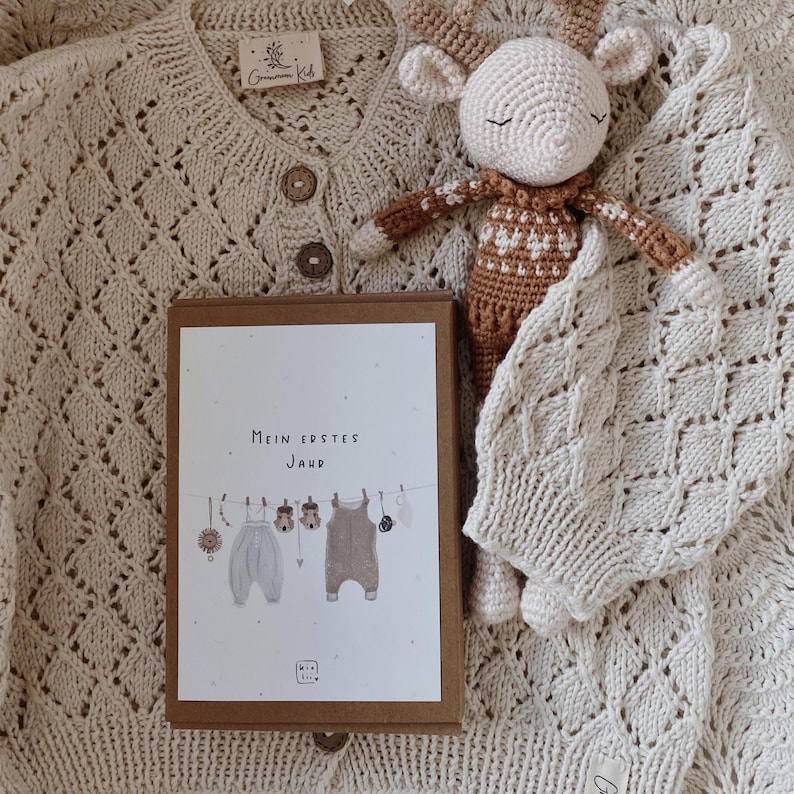 May include: A cream-colored knit cardigan with wooden buttons, a crocheted deer toy with a brown and white patterned body, and a rectangular box featuring the text "Mein erstes Jahr" with an illustration of baby clothes hanging on a line.