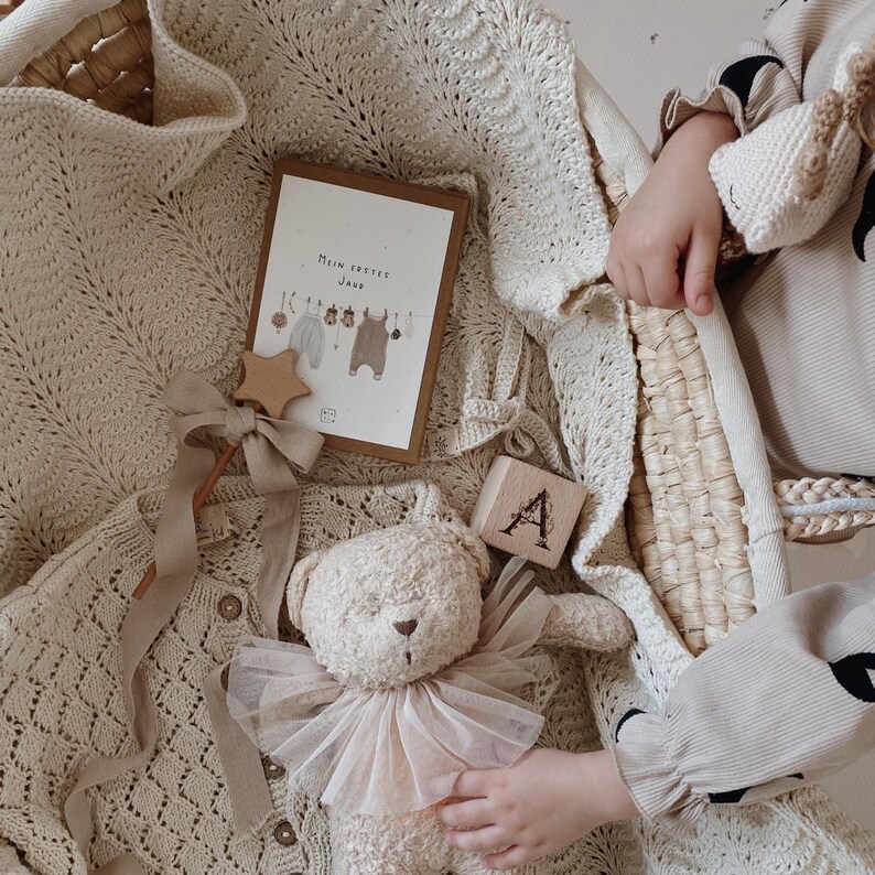 May include: A beige knitted cardigan, a teddy bear with a tulle collar, a wooden star wand with a ribbon, a wooden block with the letter A, and a rectangular card with the text Mein erstes Jahr featuring illustrations of baby clothes on a line.