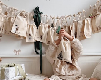 May include: A Christmas advent calendar featuring a series of small, off-white fabric drawstring bags suspended from a rope. Each bag is printed with a unique holiday-themed graphic and a number, including the number 24 on the bag held in the center.