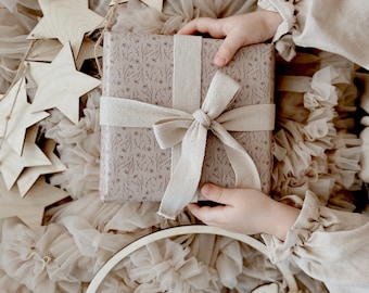 Könnte beinhalten: Eine quadratische Geschenkbox, eingewickelt in hellbeiges Papier mit einem zarten, dunkelbraunen Blumenmuster als Strichzeichnung. Die Box ist mit einem breiten, strukturierten cremefarbenen Stoffband zu einer Schleife gebunden.
