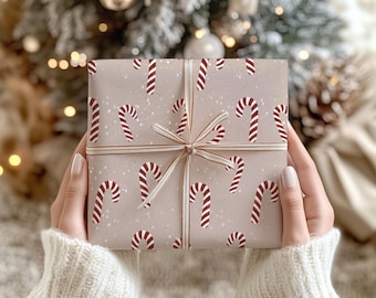 Puede incluir: Una caja de regalo cuadrada envuelta en papel marrón claro con un patrón repetitivo de bastones de caramelo rojos y blancos y pequeños puntos blancos. El paquete está atado con una cinta dorada metálica fina dispuesta en cruz con un lazo.