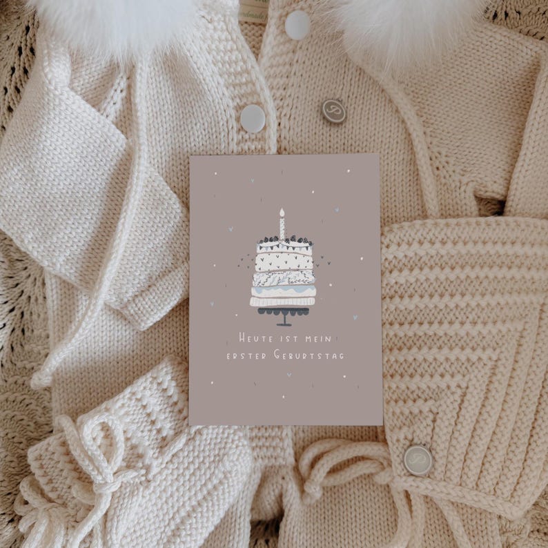 May include: A rectangular greeting card with a dusty rose background featuring a white cake illustration with a single lit candle and the text "Heute ist mein erster Geburtstag" placed over a cream-colored knitted baby outfit with a fur-trimmed hood.