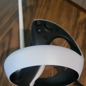 Puede incluir: Un auricular VR blanco y negro con una diadema blanca y un mando negro. El mando tiene un bot&oacute;n blanco y un bot&oacute;n X blanco.
