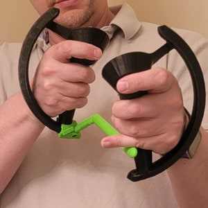 May include: A person holding two black 3D printed controllers with green handles. The controllers have a circular shape and are designed for gaming.