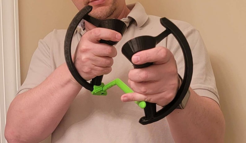 May include: A person holding two black 3D printed controllers with green handles. The controllers have a circular shape and are designed for gaming.