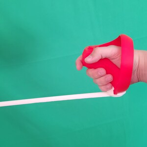 Puede incluir: Una f&eacute;rula de mano roja con una correa blanca. La f&eacute;rula est&aacute; dise&ntilde;ada para ayudar con la rehabilitaci&oacute;n de la mano.
