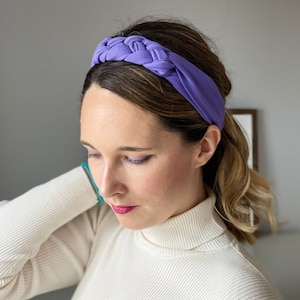 May include: A purple braided headband made of soft fabric. The headband is worn on a person's head, with their hair pulled back.