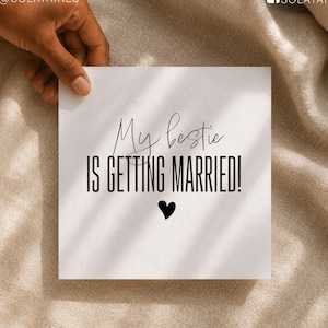 Op de afbeelding: Een vierkante witte kaart met de zwarte tekst My bestie IS GETTING MARRIED! en een klein zwart hartpictogram in het midden onder de woorden. De kaart wordt vastgehouden tegen een neutrale stoffen achtergrond met zachte schaduwen.