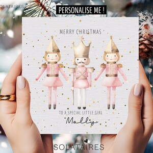 Op de afbeelding: Een kerstkaart met drie aquarel notenkrakerfiguren in roze en goud. De kaart leest "Merry Christmas" en "To a special little girl" met een naam eronder. Gouden sterren zijn verspreid over de kaart.