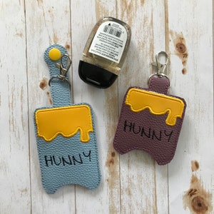 May include: Two keychains with a honey pot design. The keychains are made of faux leather and have a yellow honey pot design with the word "Hunny" written on them. One keychain is blue and the other is purple.