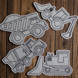 May include: Four gray construction vehicle patches with black stitching on a wooden background. The patches include a dump truck, excavator, cement mixer, and bulldozer. Each patch is outlined with black thread.