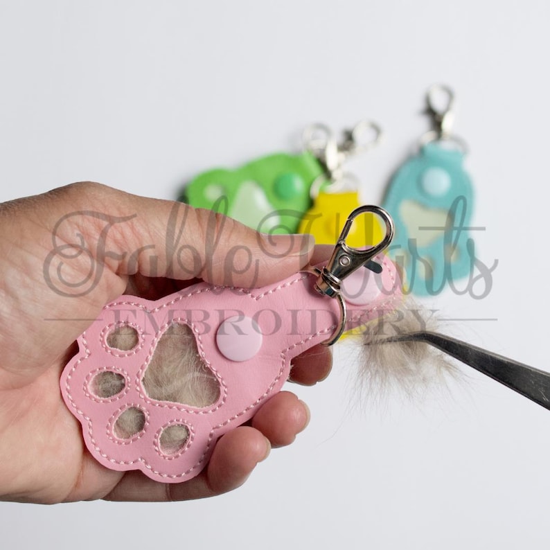 May include: A pink paw-shaped keychain with a silver clasp and a fluffy tail. The keychain has a button and stitched details. Other keychains in green, yellow, and blue are visible in the background. The text "Fable Arts Embroidery" is also visible.