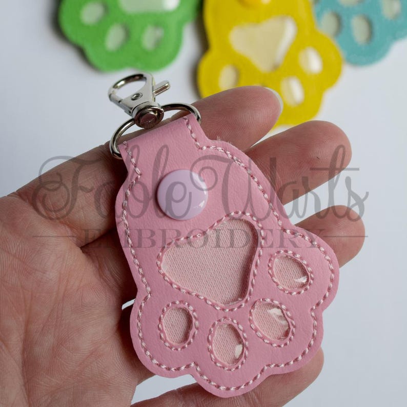 May include: A pink paw print keychain with a silver clasp and a light pink button. The keychain has a heart-shaped cutout and is stitched with white thread. Other paw print keychains in green, yellow, and blue are visible in the background.